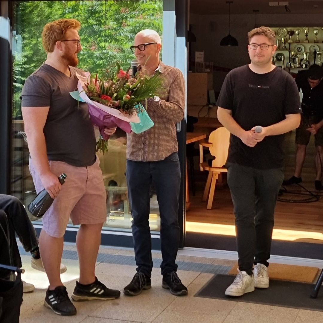Tobias Strassl bekommt von Holger einen Blumenstrauss überreicht, und Chrissi steht daneben